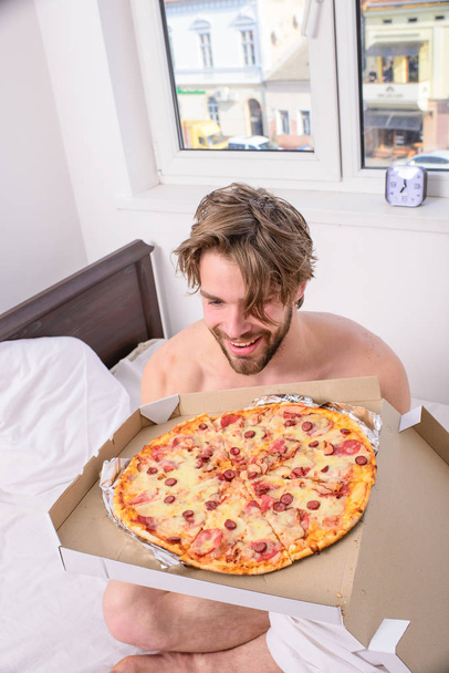 Man bearded handsome bachelor eating cheesy food for breakfast in bed. Man likes pizza for breakfast. Break diet concept. Guy holds pizza box sit bed in bedroom or hotel room. Food delivery service - Fotoğraf, Görsel