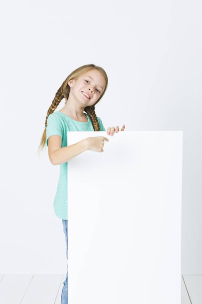 happy girl with braids in turquoise t-shirt smiling and pointing at empty board isolated on white background - Фото, изображение