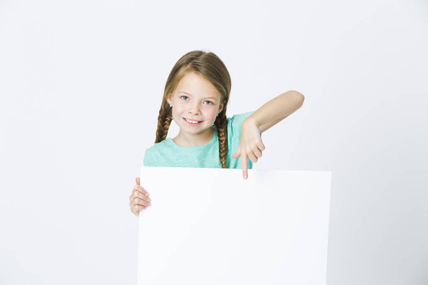 pretty little girl with braids wearing turquoise t-shirt pointing at empty board in front of white background  - 写真・画像