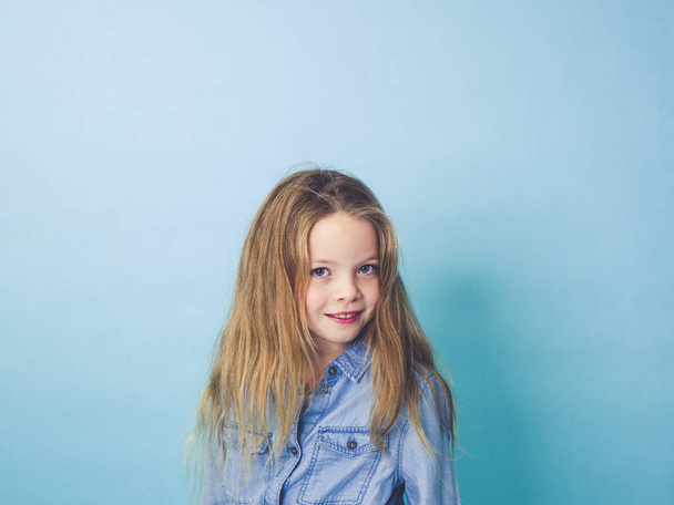 beautiful little girl looking at camera and smiling while posing in front of blue studio background  - Фото, изображение