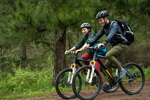 Side view of young Asian couple wearing helmets while practicing cross-country cycling on bicycles - Photo, Image