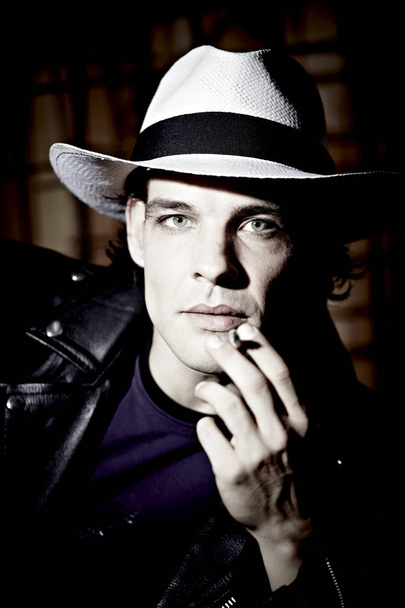 portrait of handsome young man in hat smoking cigarette on dark studio background  - Photo, Image