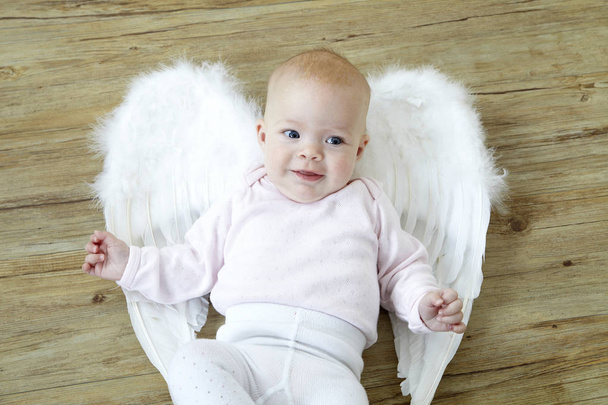 Маленький ребенок в белой одежде с ангельскими крыльями лежащими на спине и улыбающимися на деревянном полу
  - Фото, изображение