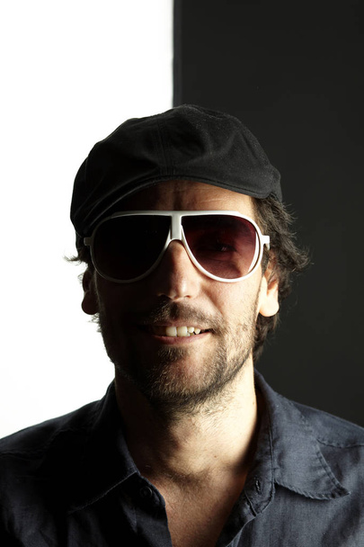 portrait of handsome man in dark cap and sunglasses smiling and looking at camera on black and white background, close-up  - Fotografie, Obrázek