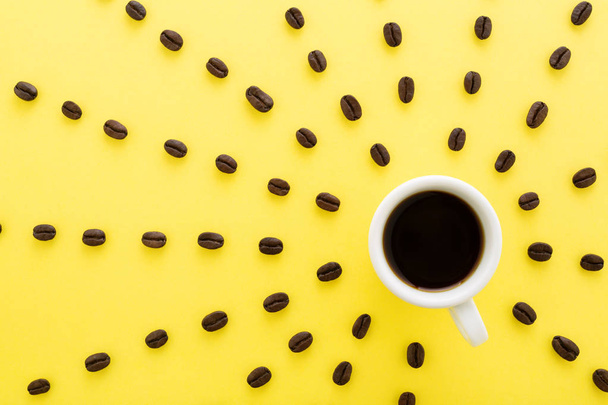 Cup of coffee espresso with sun rays of coffee beans on yellow background. Flat lay, creative design. Sunny morning & black coffee concept. - Photo, image