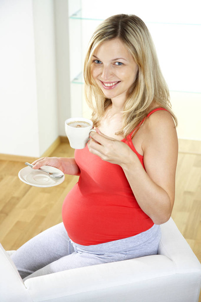happy pregnant woman sitting on armchair and drinking cofee from white cup at home - Foto, Imagem