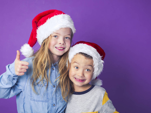 brother and sister wearing red Santa hats in front of violet background  - Foto, Bild