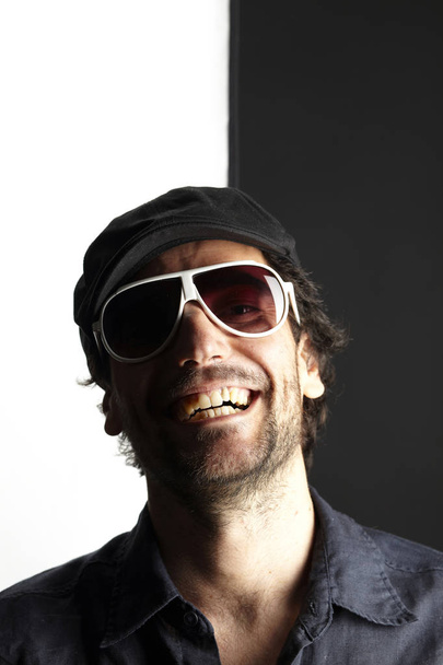portrait of funny handsome man in white cap and sunglasses laughing and looking at camera on black and white background, close-up  - Фото, изображение