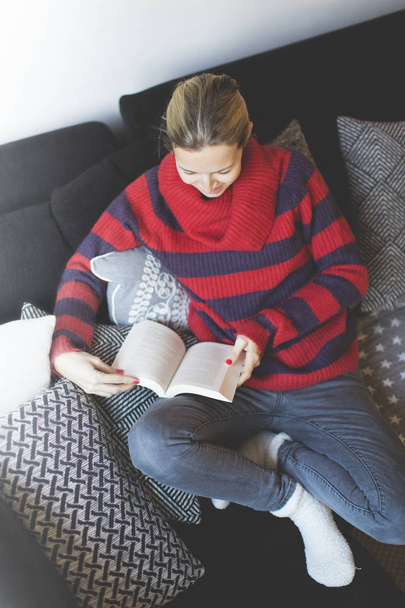 woman in red striped pullover reading a book while sitting on couch at home  - Foto, immagini