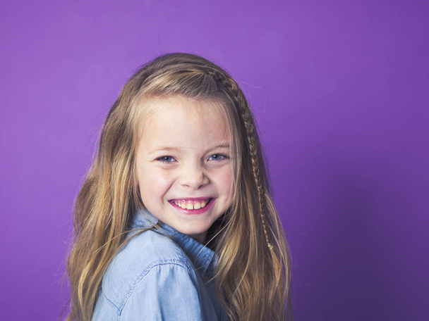 portrait of cute little girl wearing blue denim shirt in front of violet background in studio - Фото, зображення