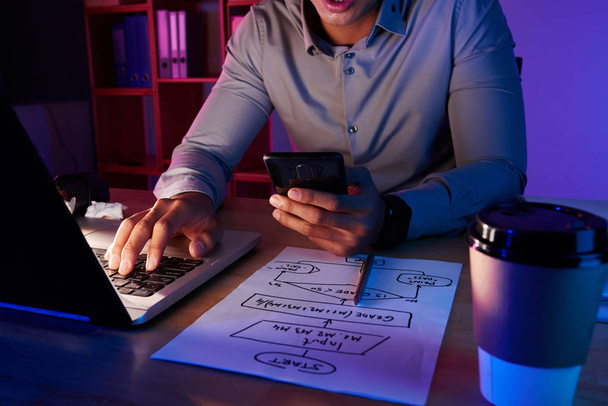 Hombre trabajador irreconocible sentado en el escritorio, marcando el número en el móvil y utilizando el ordenador portátil
  - Foto, imagen