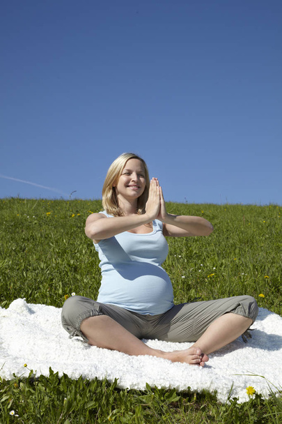 happy pregnant woman sitting on carpet and doing yoga on meadow at sunny day - Photo, Image
