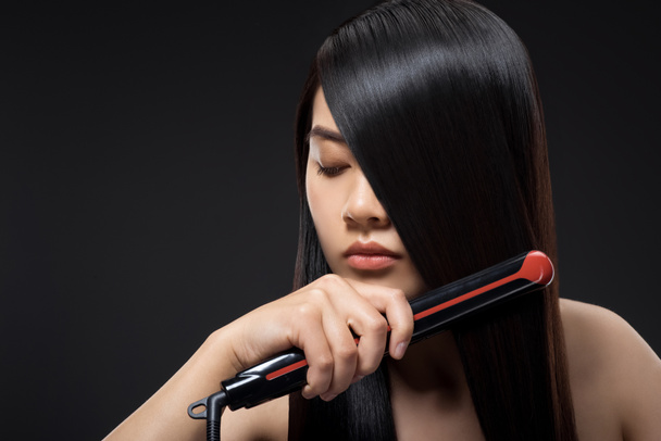 portrait of young asian woman straightening hair with hair straightener isolated on black - Photo, Image