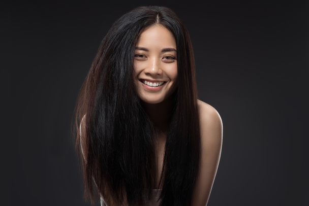 Porträt einer glücklichen asiatischen Frau mit schönen und gesunden dunklen Haaren, die isoliert auf schwarz in die Kamera blickt - Foto, Bild