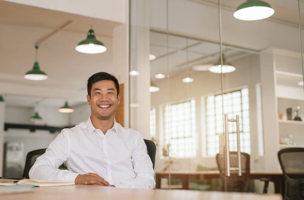Portrait of a young Asian businessman smiling confidently while sitting alone at a table in a modern office going over paperwork - Photo, Image