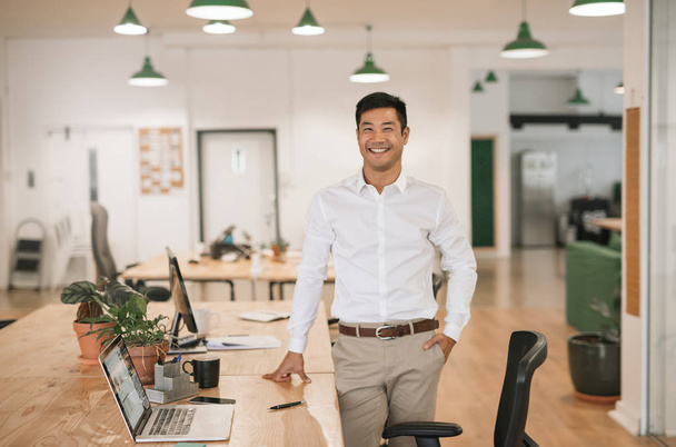 Successfull young Asian businessman smiling confidently while standing alone by his desk in a large modern office - Zdjęcie, obraz