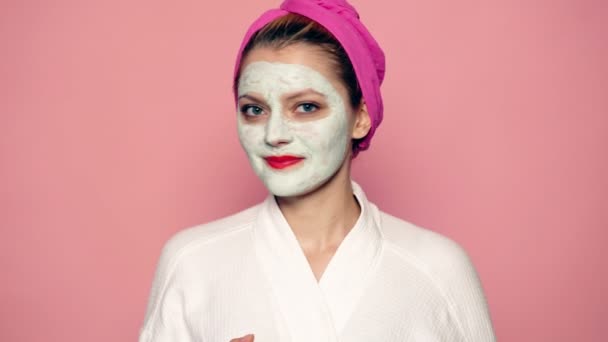 Girl with a mask on her face and a towel on her head toss up an apple on a pink background. Beautiful girl with cosmetic mask on her face. Face care concept. - Filmagem, Vídeo