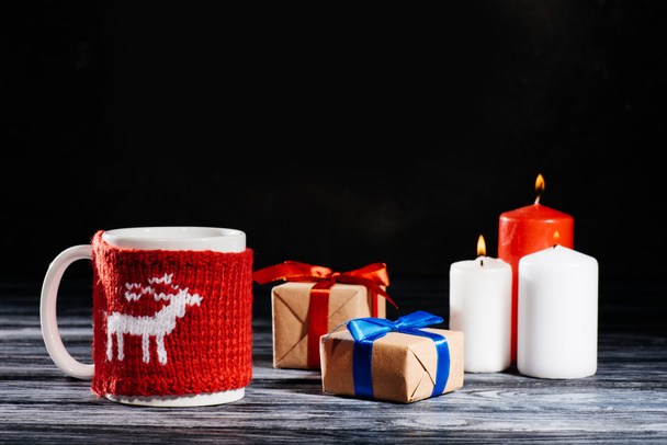 close-up view of cup, burning candles and christmas gifts on wooden table on black - Foto, Imagen