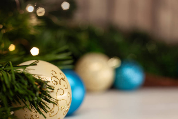 Weihnachtsspielzeug weiße Kugel mit einem glänzenden Muster neben einer blauen Kugel auf einem hellen Tisch neben einem Weihnachtsbaum auf einem Hintergrund aus Holz mit einer Girlande - Foto, Bild