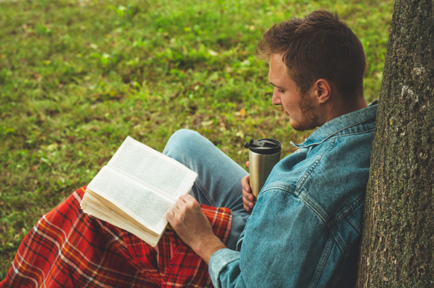 Mosolygó fiatal ember könyvével szabadtéri meleg piros kockás és egy csésze teát háttérben őszi szünet és életmód fogalma - Fotó, kép