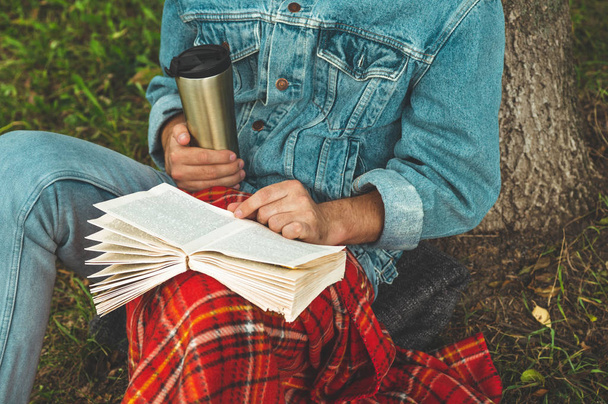 Smiling Young Man reading book outdoor with a warm red plaid and a cup of tea on background Autumn vacations and Lifestyle concept - Foto, imagen