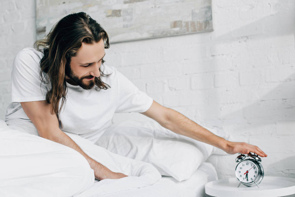 cheerful young long haired man turning off alarm clock in bedroom at home  - Photo, Image