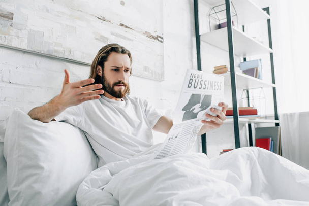 angry bearded businessman with long hair reading newspaper and gesturing by hand in bed during morning time at home - Foto, Imagem