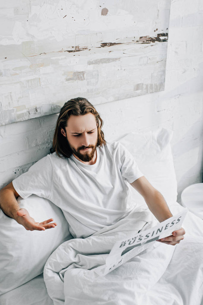 high angle view of emotional bearded businessman with long hair gesturing by hand reading newspaper in bed during morning time at home - Φωτογραφία, εικόνα