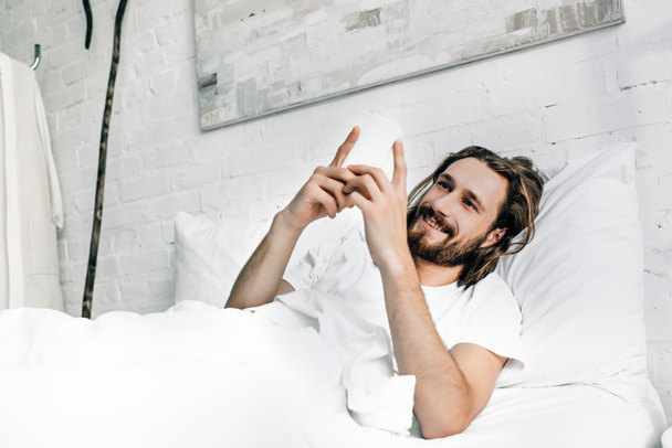 χαμογελαστός ο Ιησούς χρησιμοποιώντας το smartphone στο κρεβάτι κατά τη διάρκεια του χρόνου το πρωί στο σπίτι - Φωτογραφία, εικόνα