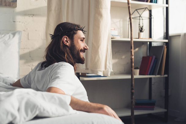 vista lateral de Jesús sonriente sentado en el suelo cerca de la cama durante la mañana en el dormitorio en casa
  - Foto, Imagen