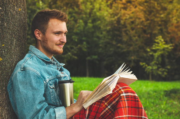 背景秋休暇とライフ スタイルのコンセプトに温かみのある赤のチェック柄と紅茶のカップで屋外の本を読んで若い男の笑みを浮かべてください。 - 写真・画像