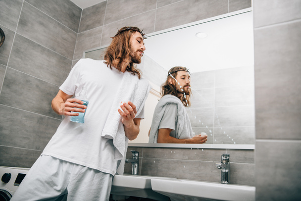 low angle view of Jesus in crown of thorns rinsing mouth by mouthwash liquid in bathroom - Фото, зображення
