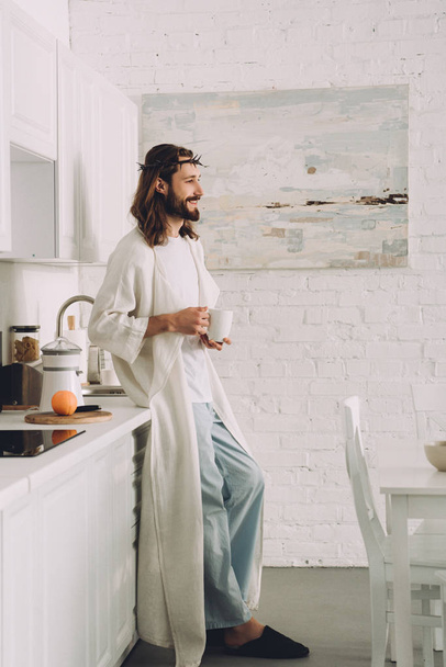 vista lateral de Jesús feliz en corona de espinas de pie con taza de café en la cocina en casa
  - Foto, Imagen