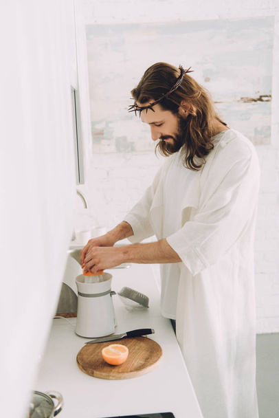 side view of cheerful Jesus doing orange juice by juicer in kitchen at home  - Fotó, kép