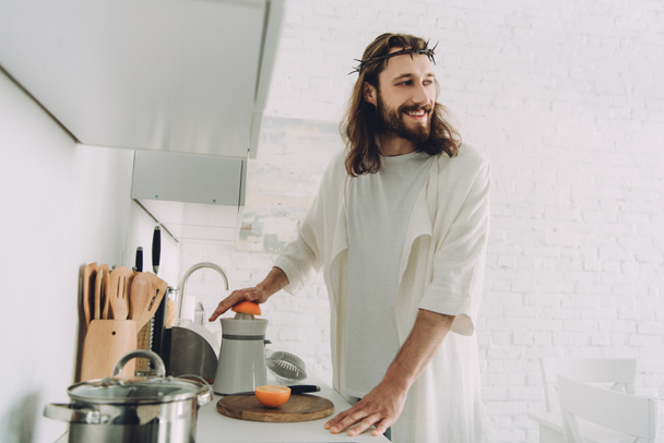 happy Jesus looking away and doing orange juice by juicer in kitchen at home  - 写真・画像