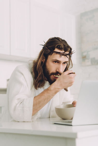 selective focus of Jesus eating corn flakes on breakfast at table with laptop in kitchen at home - Фото, зображення