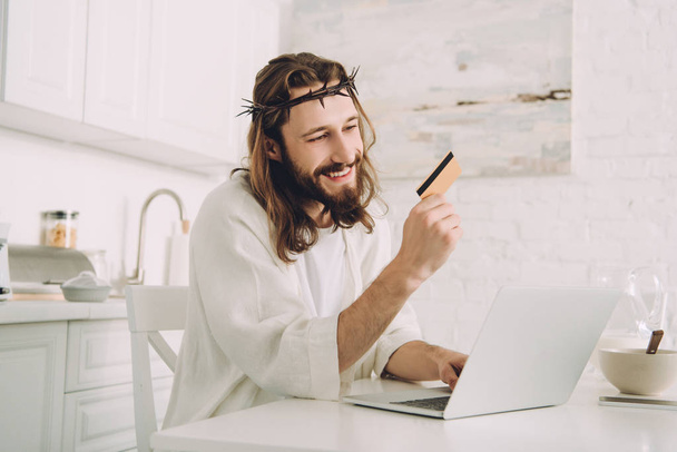 smiling Jesus holding credit card and doing online shopping at table with laptop in kitchen at home - Photo, Image