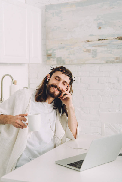vidám Jézus a fülhallgató zene és gazdaság csésze kávé asztal, laptop konyha otthon - Fotó, kép