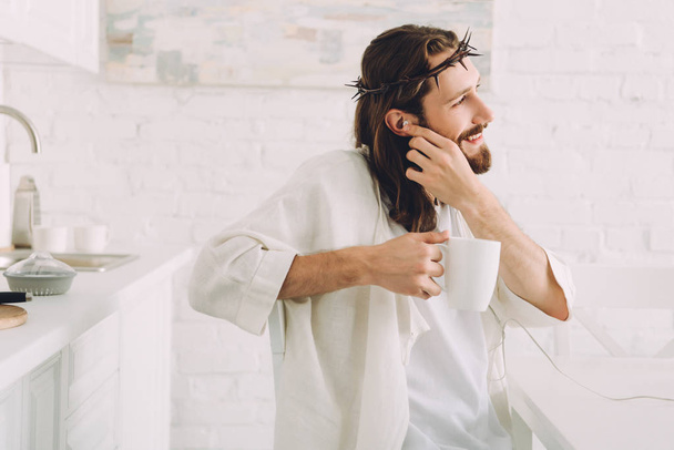 side view of happy Jesus in earphones listening music and holding cup of coffee in kitchen at home - Photo, Image