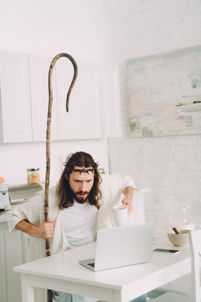 shocked Jesus with coffee cup and wooden staff looking at laptop in kitchen at home  - Zdjęcie, obraz