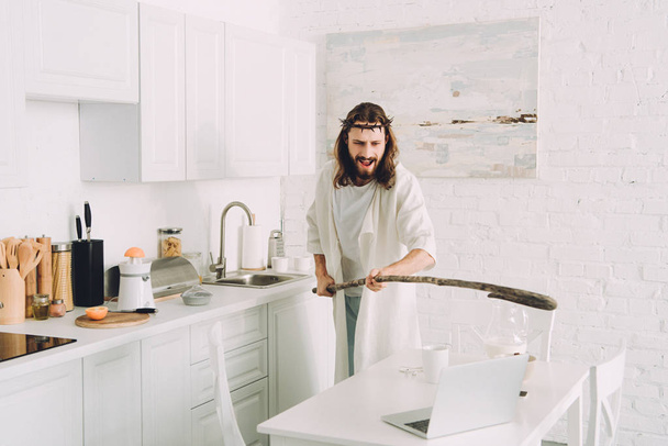 aggressive Jesus hitting laptop by wooden staff in kitchen at home  - Photo, Image