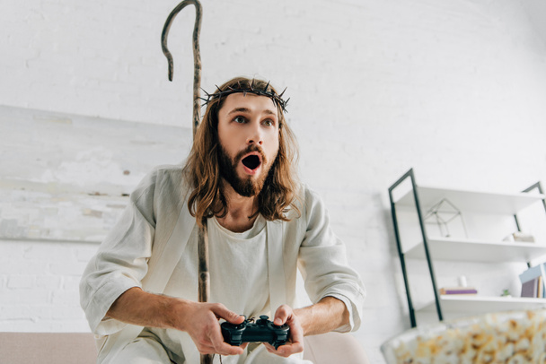 shocked Jesus with wooden staff playing video game by joystick on sofa at home - Photo, Image