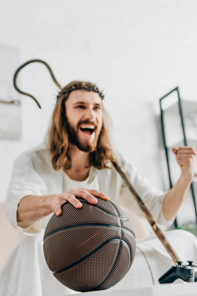 excited Jesus with wooden staff gesturing by hand and watching basketball match on sofa at home - Фото, изображение