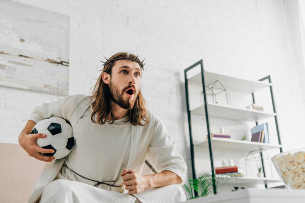 Jesús emocional en corona de espinas sentado en el sofá con pelota de fútbol y viendo el partido de fútbol en casa
 - Foto, imagen