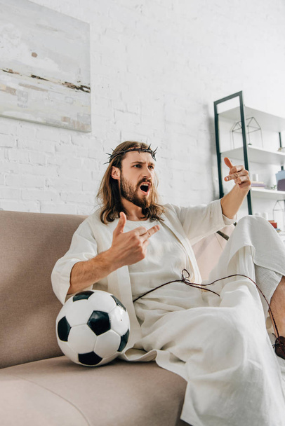 aggressive Jesus in crown of thorns watching football and showing middle fingers match at home - Photo, image