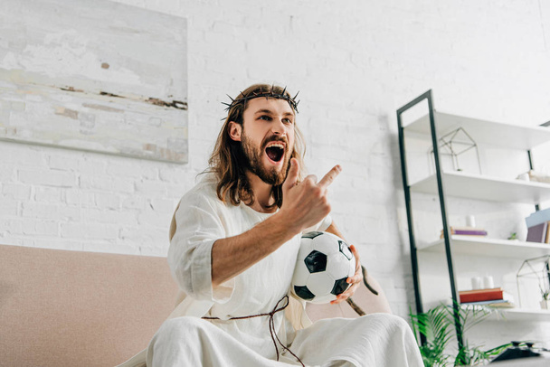angry Jesus in crown of thorns showing middle finger during watching of soccer match at home - Photo, Image