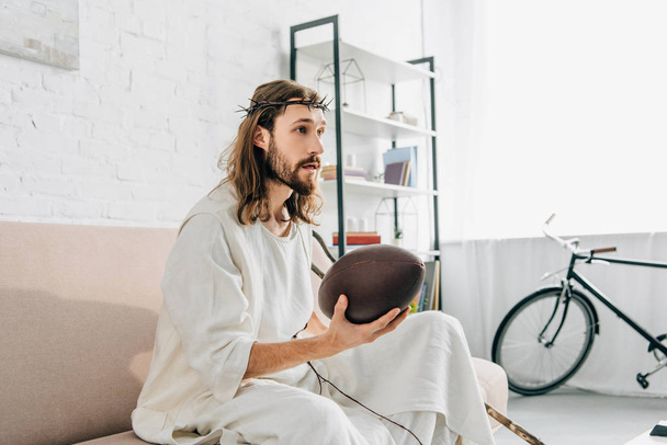selective focus of Jesus in crown of thorns holding ball and watching rugby on sofa at home - Фото, изображение