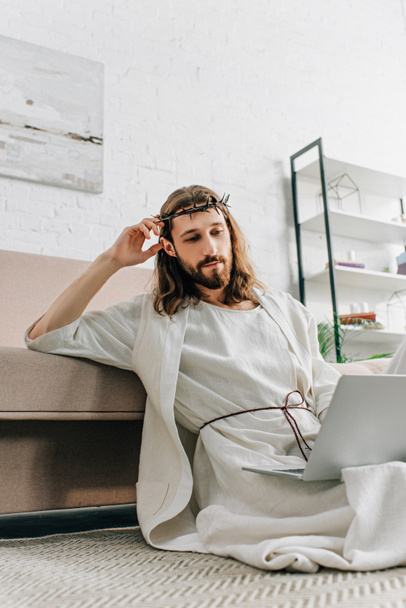 confident Jesus in crown of thorns sitting on floor and using laptop near sofa at home - 写真・画像