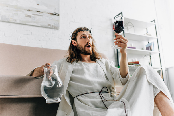 shocked Jesus in crowns of thorns and robe holding jug of water and looking at wine glass in hand at home - Φωτογραφία, εικόνα