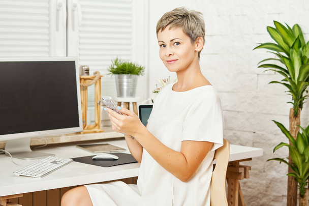 bela mulher de negócios em um vestido branco sentado em uma mesa com um telefone na mão e trabalha no computador em um escritório em casa
 - Foto, Imagem